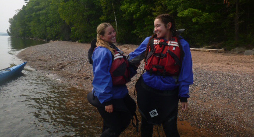 two women wearing life jackets stand on the shore of a lake in ankle deep water, laughing together